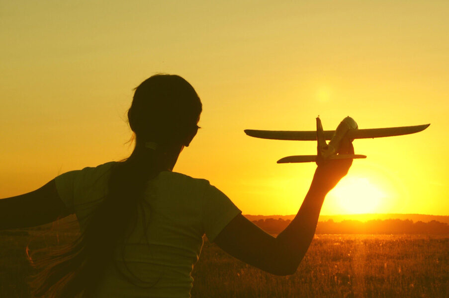 A teen or tween girl holding a model airplane as if it is flying into the sunset sky