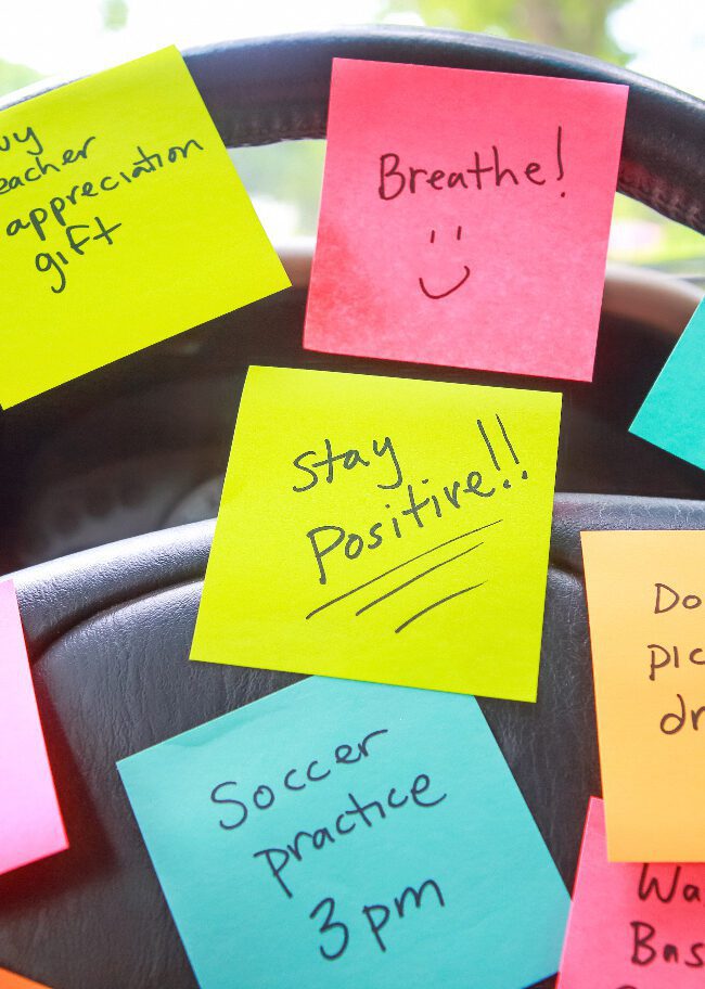 Post It notes stuck to the steering wheel of a car reminding a parent to pick up dinner, drive to soccer practice, and "Stay positive!"