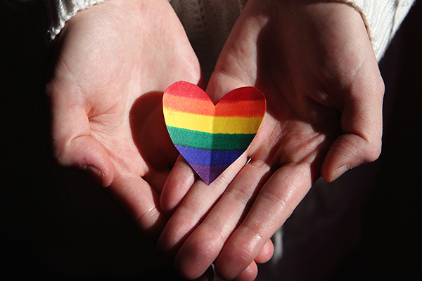 A paper heart with rainbow stripes held lovingly in cupped open hands to signify support for the LGBTQ community