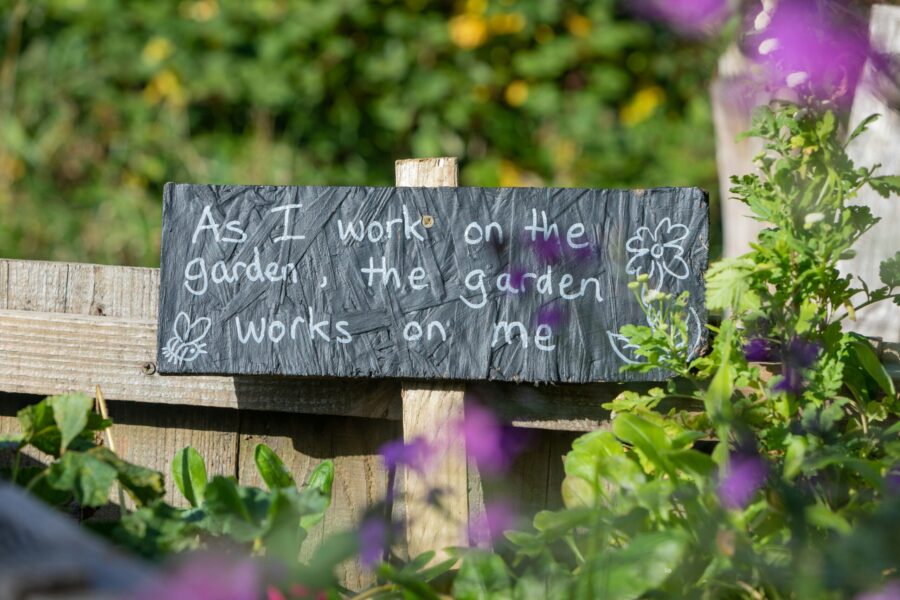 A sign in a garden which reads, "As I work in the garden, the garden works on me"