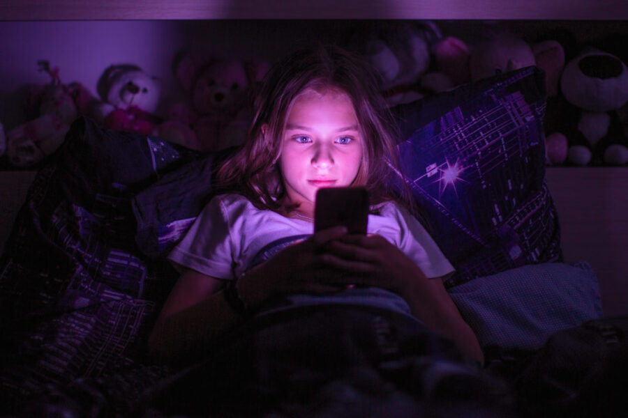 A tween girl up late at night, her face lit by the glow of her smartphone